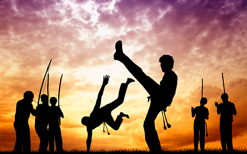 Em um fim de tarde, sete pessoas aparecem jogando capoeira.